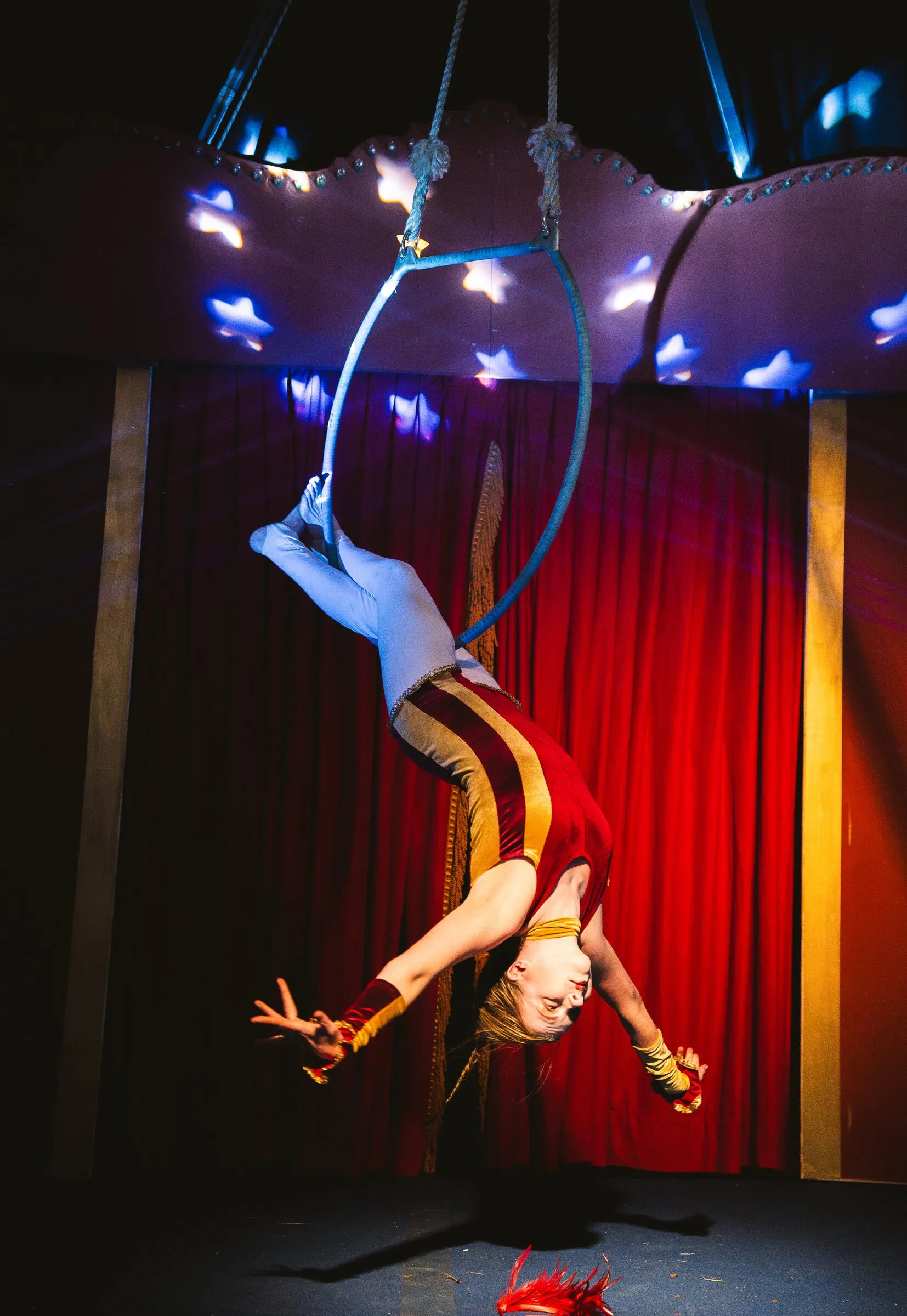 A Kuroneko Circus aerialist hanging backward from an aerial hoop beneath a canopy of lit stars