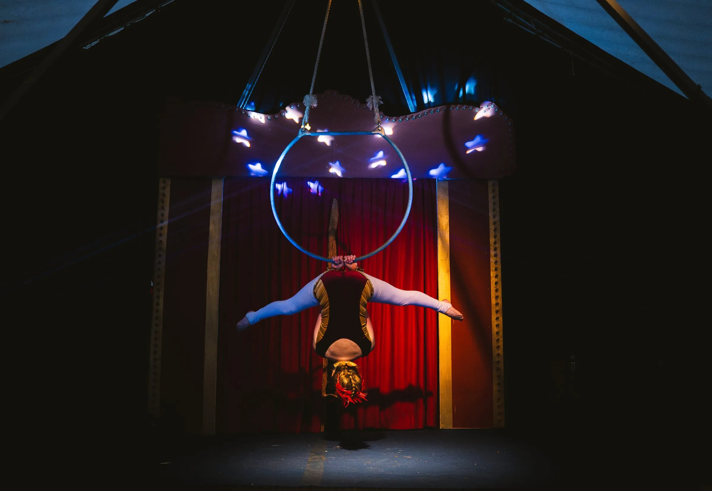A Kuroneko Circus aerialist in an inverted split beneath an aerial hoop under the lit star canopy