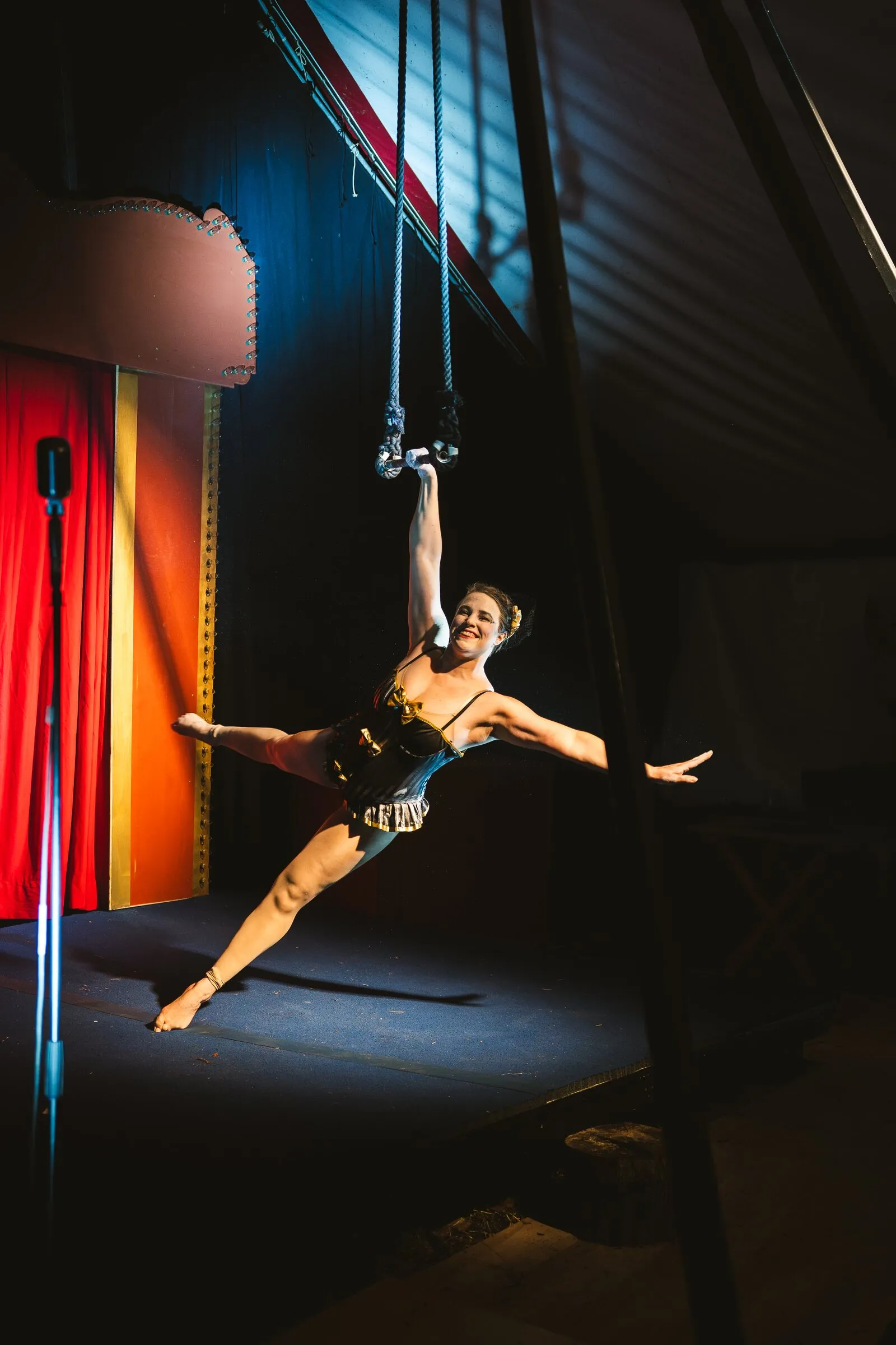 A Kuroneko Circus aerialist hanging from one arm on aerial straps, legs split in a spotlight