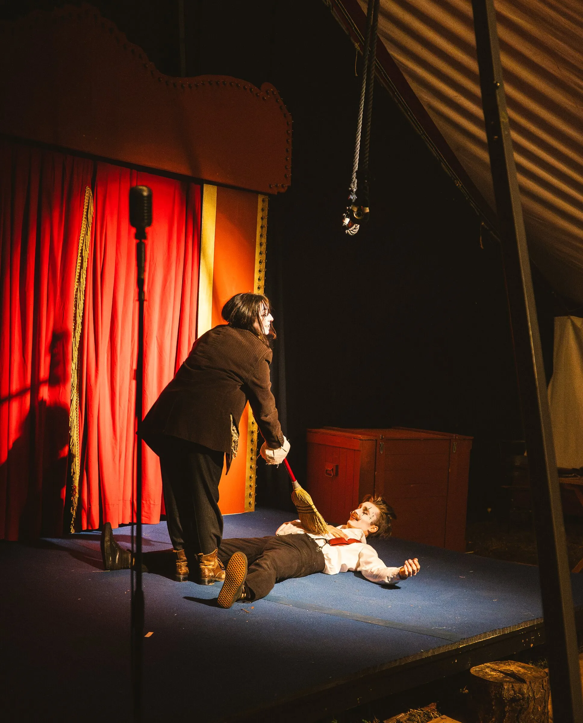 A Kuroneko Circus clown sweeping a broom over a partner lying flat on the stage