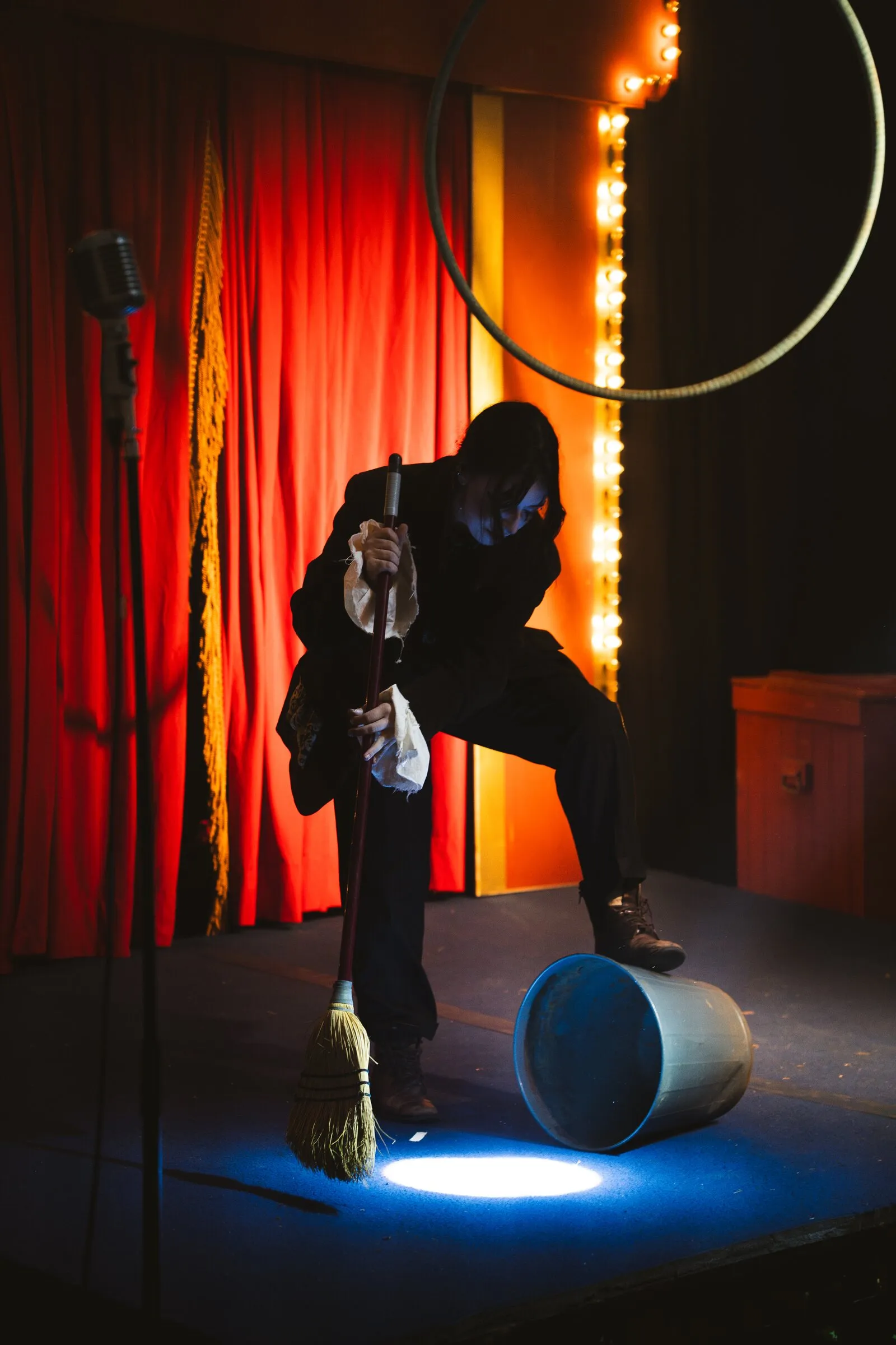 A Kuroneko Circus clown with a broom and an overturned bucket caught in a pool of spotlight
