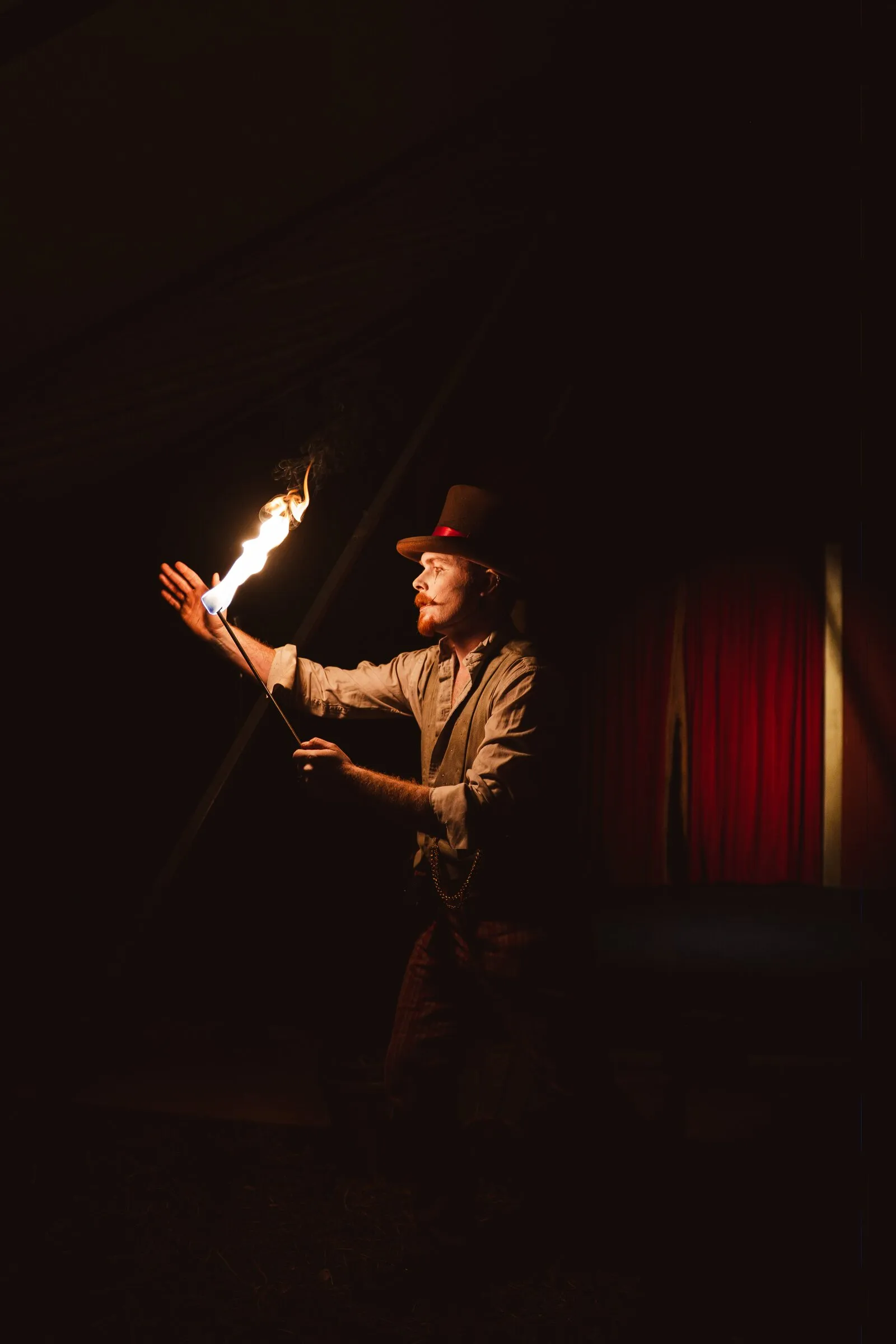 A Kuroneko Circus fire performer in a top hat raising a lit torch in the dark tent