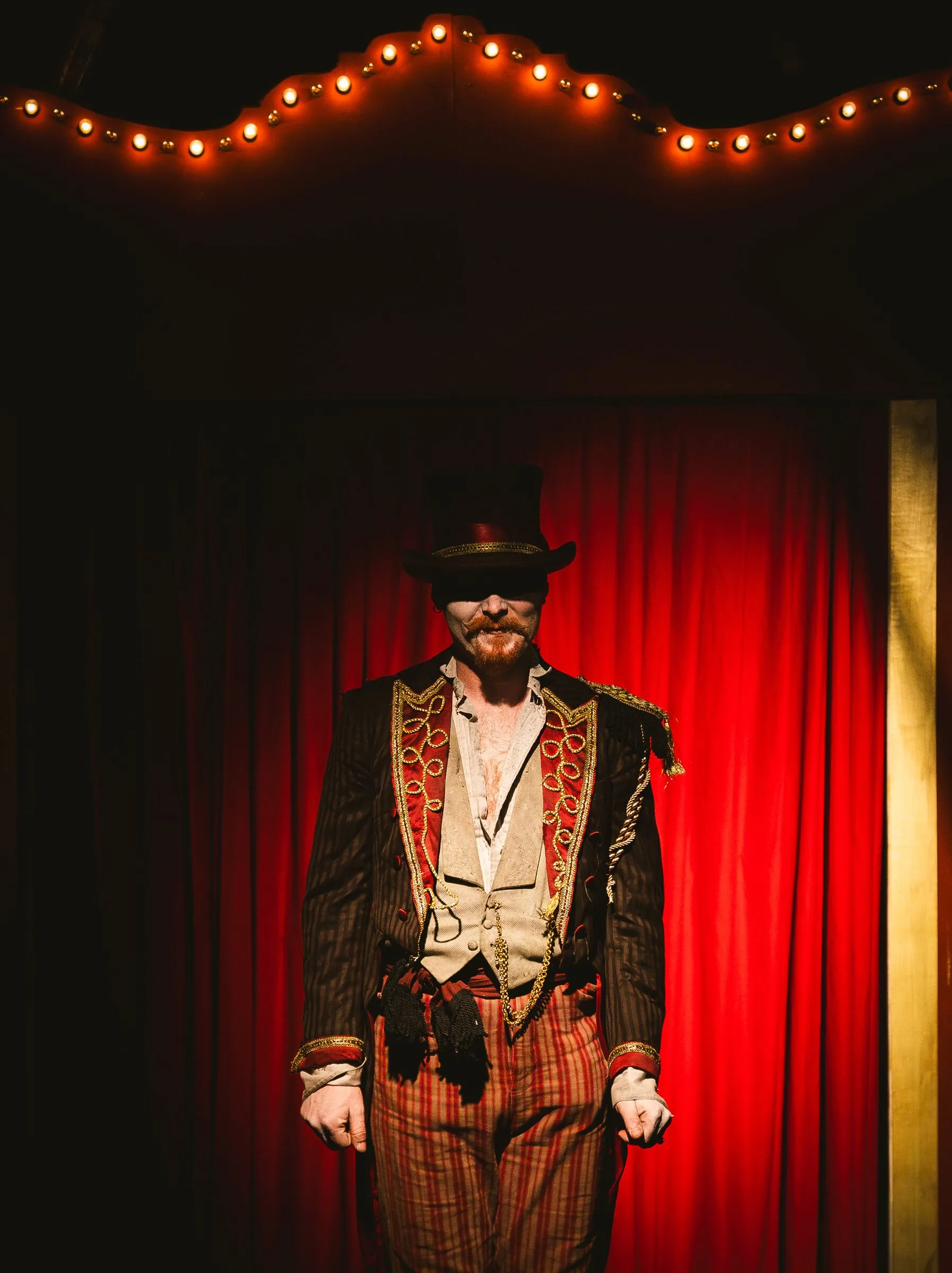 The Kuroneko Circus ringmaster in a top hat and braided coat standing before the red curtain