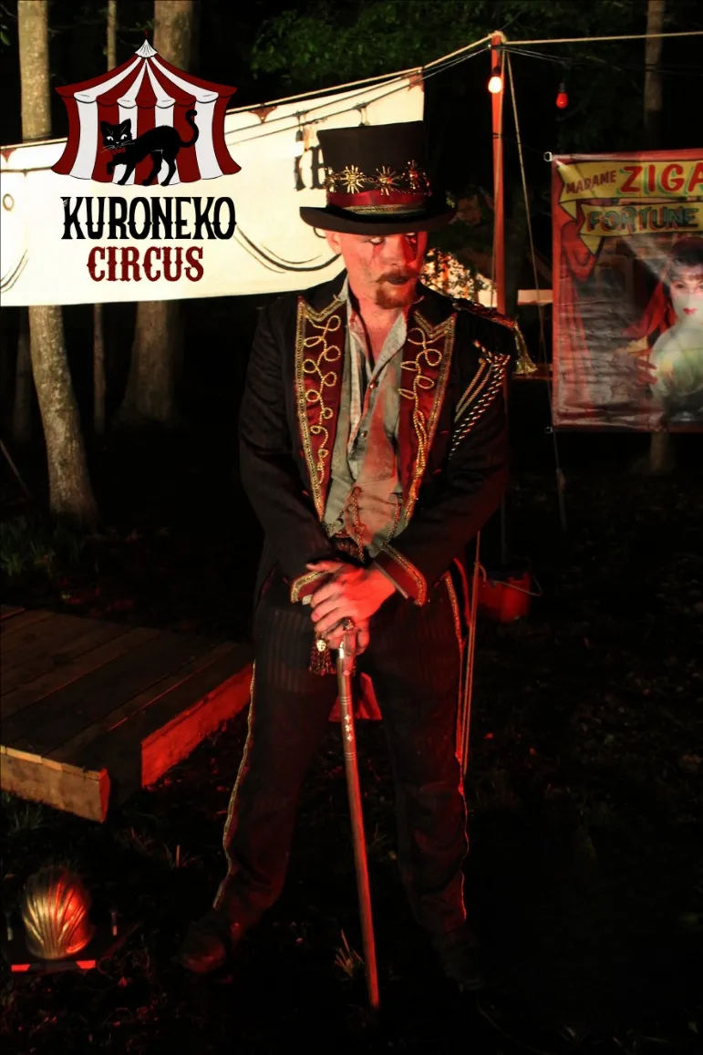 A Kuroneko Circus ringmaster in an ornate coat leaning on his cane beside the fortune teller's banner during a nighttime North Carolina performance