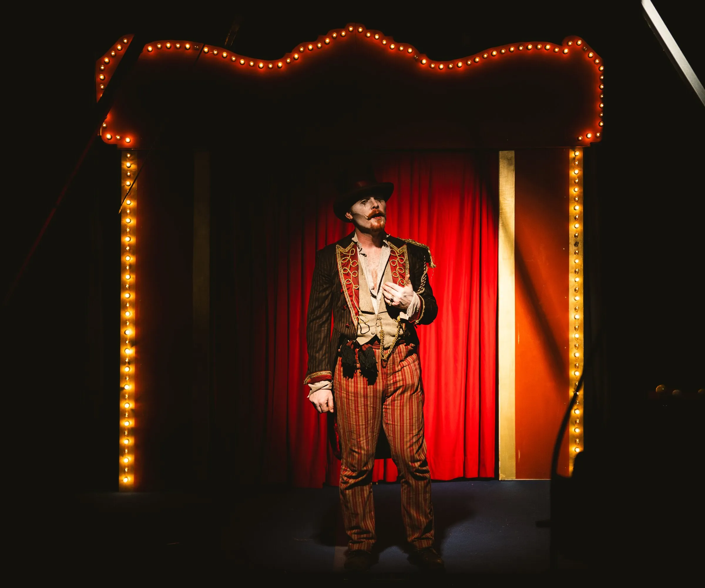 The Kuroneko Circus ringmaster in a braided coat standing center stage beneath the marquee lights