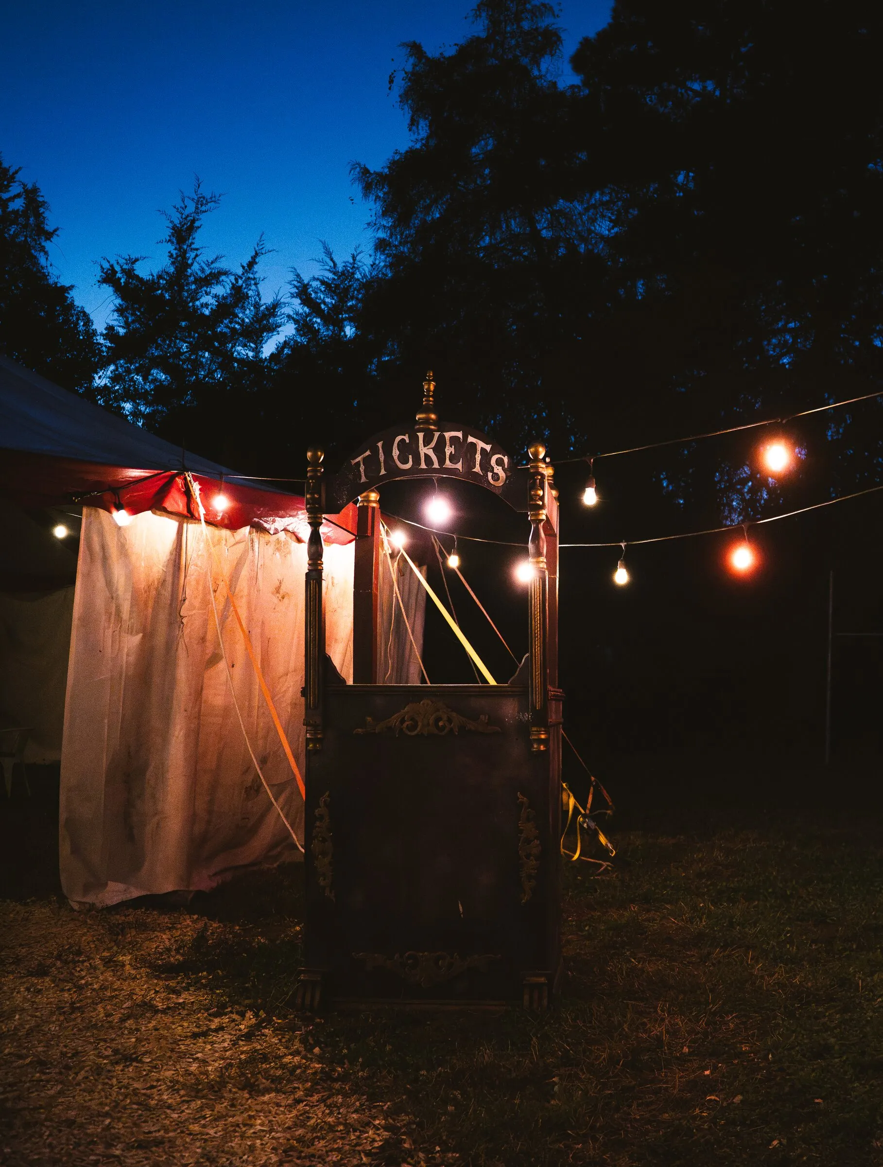 The vintage Tickets booth beside the Kuroneko Circus tent under string lights and a deep blue sky